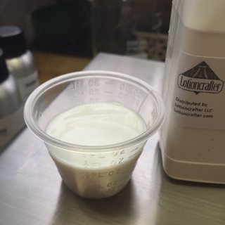 A shot glass filled with a white, opaque, thicker liquid stands beside a bottle. The liquid is Sepiplus 400, a cold process liquid emulsifier.