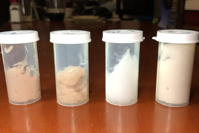 Four vials of lotion sit on a table. The first two have a brown/beige colour. The third is almost a bright white. The fourth is a lighter beige colour. Used to illustrate the impact of vanilla on a formula.