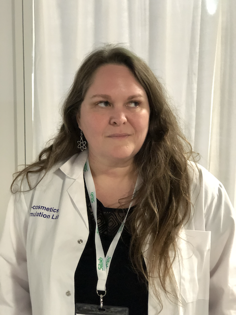 A middle aged, whites woman with linger brown hair stands in front of a white curtain in a lab coat