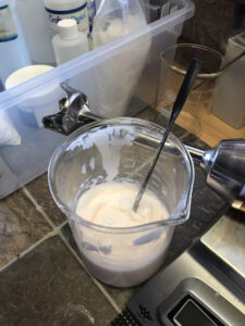 A beaker filled with conditioner sits on a countertop with a thermometer in it and an immersion blender next to it on the counter