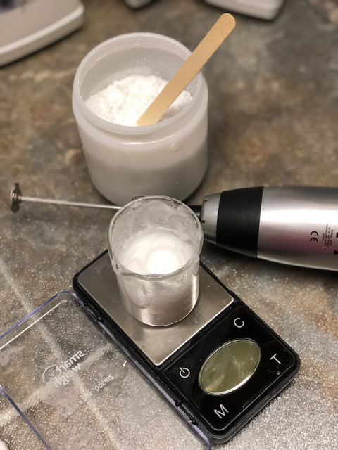 A beaker sits on a digital scale with a white lotion in it. There's a container with a powder, Penstia powder, and a hand held immersion bender beside it.
