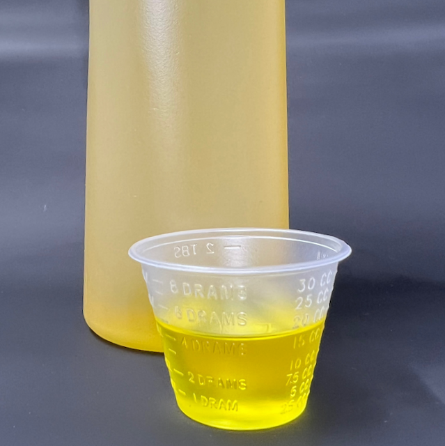 A cup of avocado oil, a thick, yellow oil. Behind it is a portion of the bottle with the oil inside.