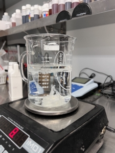 A beaker with a water and a patch of white liquid on the bottom sits on a digital scale in a lab 