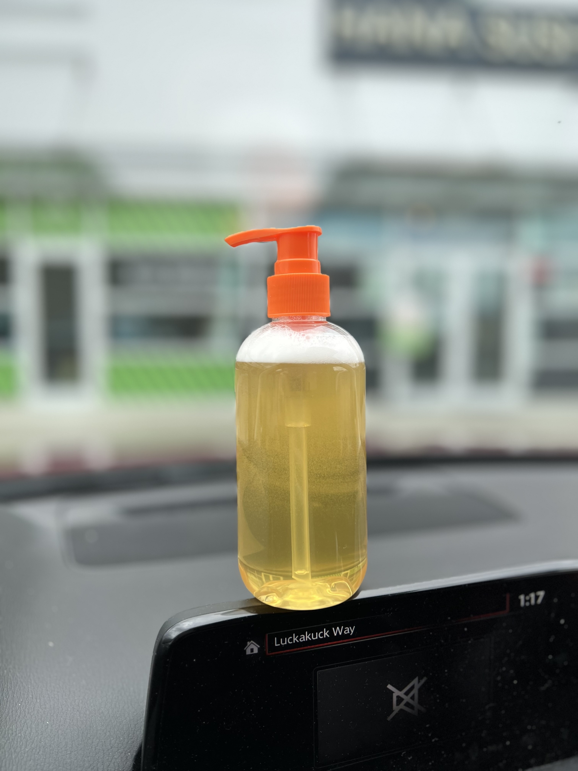 A bottle filled with a light yellow shampoo with an orange pump cap sits on a black dashboard