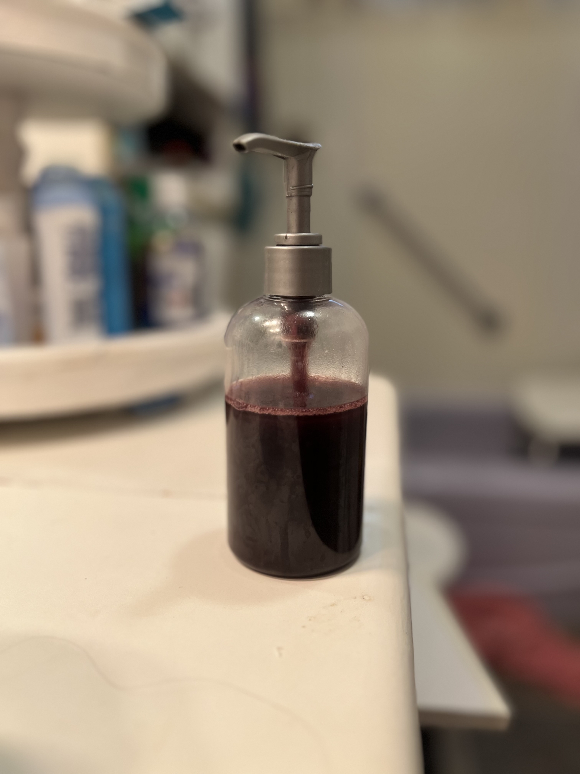 A bottle with a silver pump sits on a white counter in a bathroom. The bottle contains a red- purple liquid, a body wash with too many extracts.