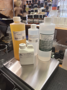 Ingredients for a Zoom workshop on a scale. Bottles of rice bran oil, Sapogel Q (NatraSap Q™), fractionated coconut oil, and squalane. 