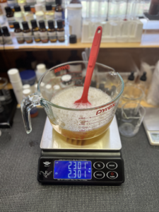 A yellow liquid in a Pyrex jug with a large fork on it on a scale with three bottles of surfactant beside it. Taken in portrait mode.