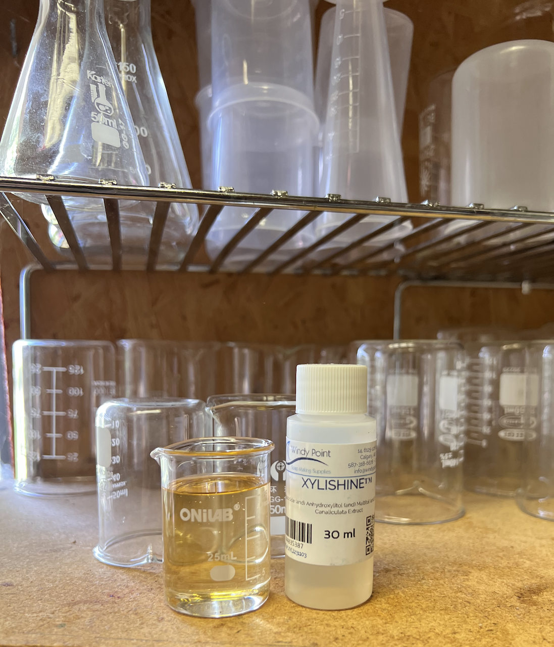 A bottle of XYLISHINE™ beside a beaker of thicker, clear, yellow liquid, which is XYLISHINE™™. They sit on a wooden shelf with a number of beakers behind them
