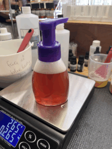 A foamer bottle sits atop a digital scale, a reddish foamy liquid inside. This is the foaming blueberry & chamomile facial cleanser, a formula for which I'll share later in July 2025