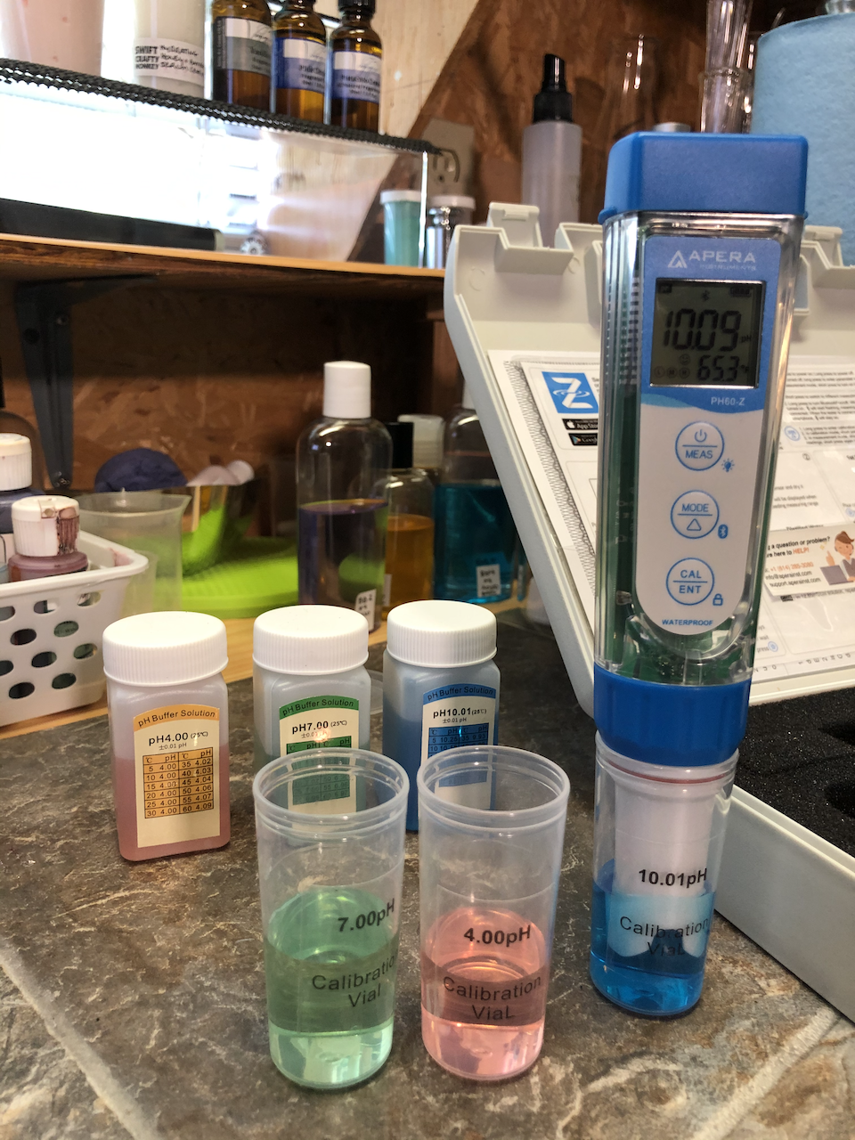 Apera pH 60 pH meter with the pH solutions and screen turned on. It's a blue pen type pH meter with an LED screen and white inset where the buttons are
