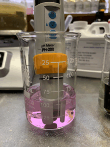 A beaker sits on a table, a white and orange pH meter is sitting in pink liquid to demonstrate how we need to cover the electrode with all the liquid