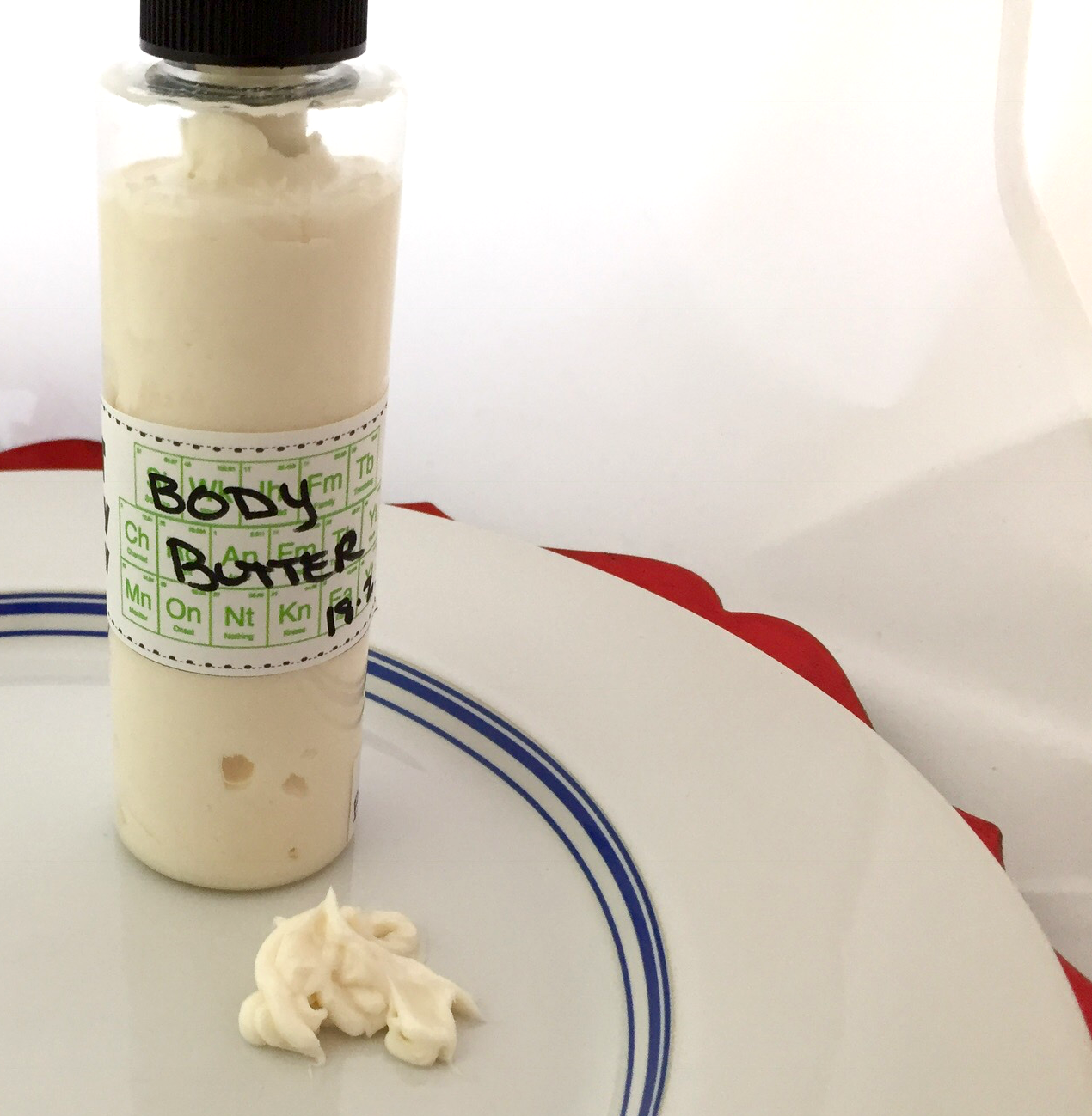 A photo of a bottle of lotion with "body butter" sitting on a white plate with a blue line. You can see the body butter is thick, rich, creamy, and off-white.